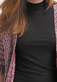 Černý žebrovaný rolák pod růžovým vzorovaným cardiganem. Cardigan má krátké rukávy a texturovanou vazbu s převážně růžovým a bílým designem.