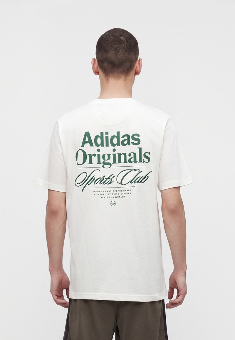 Mann, der ein weißes Adidas Originals Sports Club T-Shirt mit grünem Text trägt, steht mit dem Rücken vor einfachem Hintergrund.
