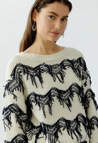 Weißer Strickpullover mit einem wellenförmigen Design in Marineblau und Fransenakzenten. Der Halsausschnitt ist weit, und die Textur ist dick und warm.