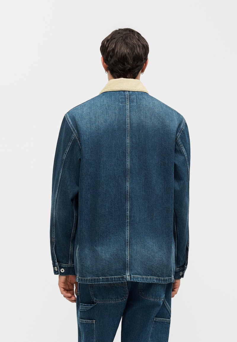 Man gezien van achteren, gekleed in een blauw spijkerjack met een beige kraag en bijpassende spijkerbroek tegen een effen achtergrond.