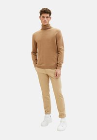 Brun polokrage tröja i mjukt stickat material, matchad med beige slim fit-byxor och vita sneakers. Modellen står framför en vit bakgrund.