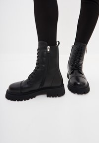 Zwarte leren enkelboots met een robuuste zool, voorzien van een zijrits, veters en textuurelementen. Gladde afwerking met een ronde neus.