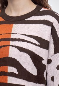 Gros plan sur un pull en maille multicolore avec des motifs abstraits marron, orange et blanc sur le haut du torse et le cou d'une personne.