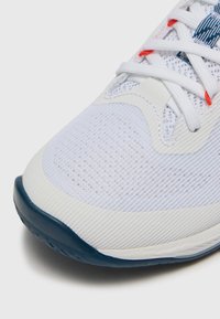 Chaussure de sport blanche avec une tige en mesh texturé, semelle en caoutchouc, accents bleus et détails rouges sur les lacets et l'étiquette.