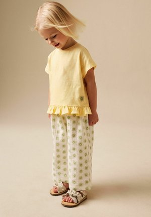 Junges Kind in gelbem Rüschentop und weißer Hose mit grünen Sonnenmustern, trägt beige Sandalen, steht vor einem einfarbigen beigen Hintergrund.