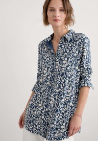 Hellblau gemustertes Hemd mit weißem Blumendesign, Knopfleiste vorne, Kragen, lange Ärmel, die an den Handgelenken hochgerollt sind, leichtes Material.
