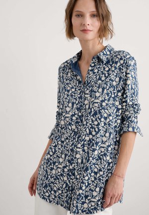 Chemise bleue à motifs avec un design floral blanc, boutonnée sur le devant, col classique, manches longues retroussées aux poignets, tissu léger.