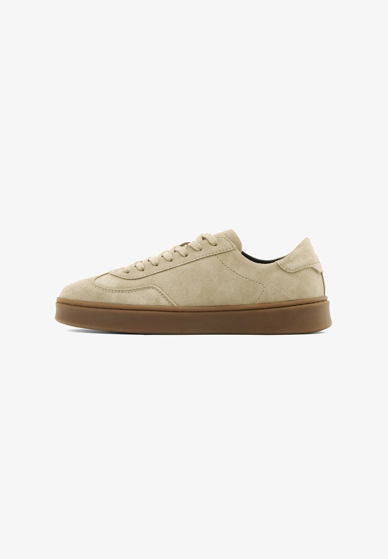 Beige suède lage sneaker met bijpassende veters en dikke bruine rubberen zool, zijaanzicht op witte achtergrond.