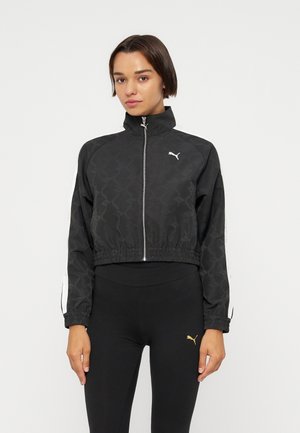 Donna che indossa una giacca da ginnastica nera Puma con zip frontale e leggings neri con un piccolo logo Puma dorato.