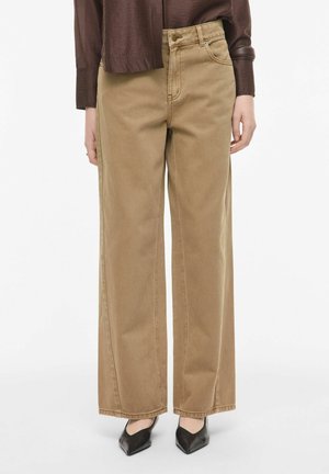 Person trägt hochtaillierte, weit geschnittene, beige Hose, dunkelbraunes langärmeliges Oberteil und schwarze spitze Schuhe, steht vor weißem Hintergrund.