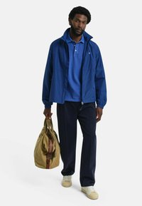 Sininen tuulenpitävä takki sinisen poolopaidan päällä, tummat housut ja kermaiset lenkkarit. Ruskea kankainen duffel-reppu, jossa on punainen raita.