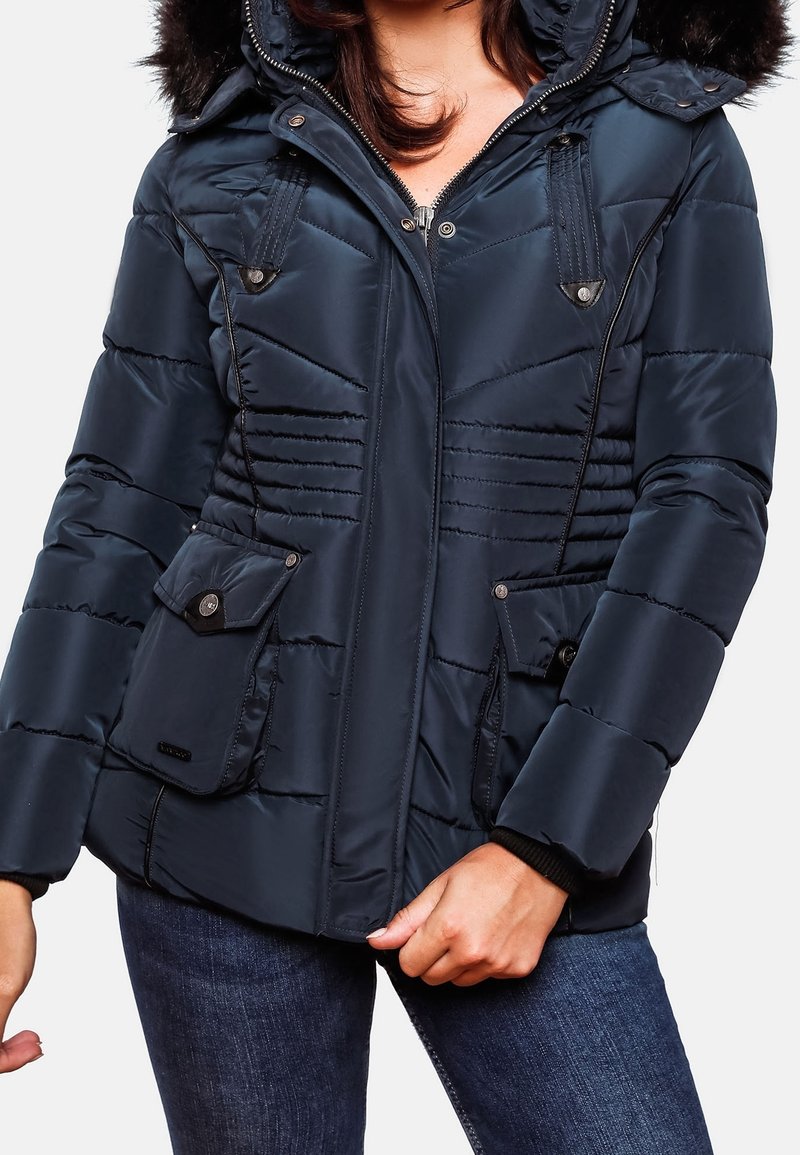 Femme portant une veste d'hiver matelassée bleu marine avec une capuche bordée de fourrure et des poches à rabat, associée à un jean bleu foncé.