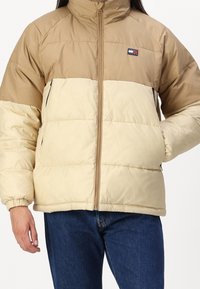 Tan and beige puffer jacket with a zip front, high collar, ribbed cuffs, and side pockets. The fabric has a textured pattern.