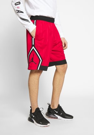 Osoba nosząca czerwono-czarne spodenki do koszykówki z logo koszykówki i czarne buty sportowe Nike stojąca na białym tle.
