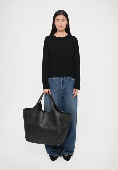 Sort læder tote-taske med stor åben top og robuste håndtag, vist i hænderne på en person iført afslappet outfit med sort sweater og blå jeans.