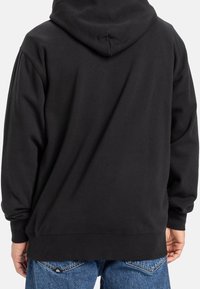 Una persona che indossa una felpa nera con cappuccio e jeans blu, vista di spalle su uno sfondo bianco.