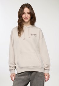 Beżowa bluza z kapturem z miękkiego materiału, posiada kieszeń z przodu, kaptur ze sznurkiem oraz haftowany napis "Dziś jest super" w kolorze brązowym. Noszona z szarymi dżinsami.