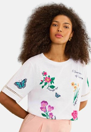 Kvinde med voluminøst krøllet hår iført en hvid T-shirt med farverige broderede blomster og sommerfugle, kombineret med lyserøde bukser.