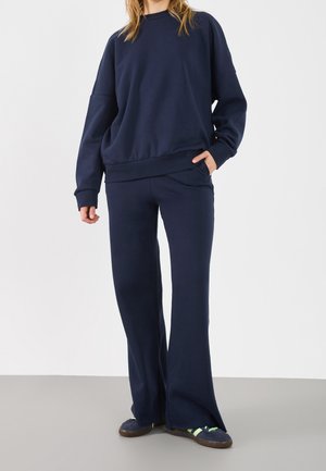 Personne portant un sweat-shirt oversized bleu marine et un pantalon large assorti avec des baskets foncées sur un fond blanc uni.