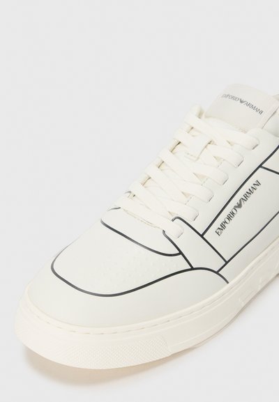 Sneaker en cuir blanc avec des contours noirs, textures perforées et un panneau latéral marqué. Comprend des lacets plats et une semelle en caoutchouc texturé.