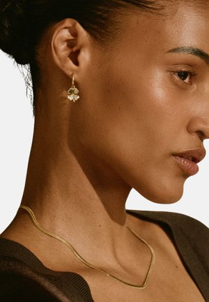 Goudkleurige bloemoorbellen met een gladde textuur en een getextureerde, gebogen gouden ketting op een model met donker haar.