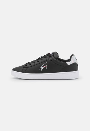 Baskets basses noires avec semelle blanche, lacets noirs et un détail métallique au talon, avec un petit logo sur le panneau latéral.