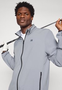 Jachetă de golf gri deschis cu accente negre, cu închidere cu fermoar, protecție pentru bărbie și manșete elasticizate, purtată de o persoană care ține o crosetă de golf.