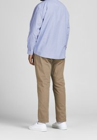 Camicia a maniche lunghe blu chiaro, pantaloni color beige e sneakers bianche; cuciture visibili, bottoni sulle maniche e una vestibilità rilassata. Vista posteriore.