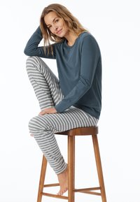 Maglietta a maniche lunghe color teal abbinata a leggings a righe grigio chiaro e bianchi, seduta su uno sgabello di legno. Tessuto morbido e traspirante.