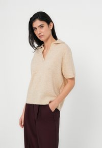 Πουλόβερ με κοντά μανίκια, μπεζ πλεκτό, με V-λαιμόκοψη και γιακά, με απαλή υφή και άνετη εφαρμογή. Συνδυάζεται με σκούρο βουργουνδί παντελόνι.