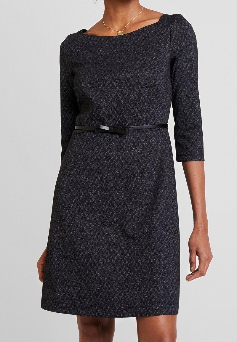 Femme portant une robe ajustée à motifs sombres avec des manches trois-quarts et une fine ceinture noire ornée d'un petit nœud à la taille.