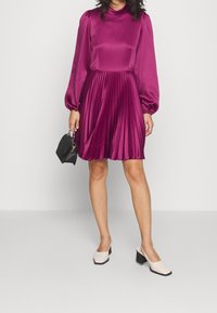 Robe en satin fuchsia avec jupe plissée, col haut et manches ballon. Associée à un sac à main noir et des chaussures à talons carrés crème.