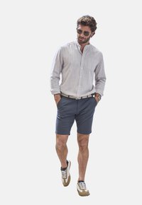 Helles gestreiftes Hemd, marineblaue Shorts und mehrfarbige Sneaker. Accessoires sind Sonnenbrille und ein gewebter Gürtel. Lässige Kleidung.
