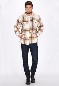 Ruudullinen flanellitakki vaaleassa ja ruskeassa, jossa on pehmeä beige sherpa-kaulus, kaksi rintataskua ja napitus edessä. Yhdistetty tummiin farkkuihin ja saappaisiin.