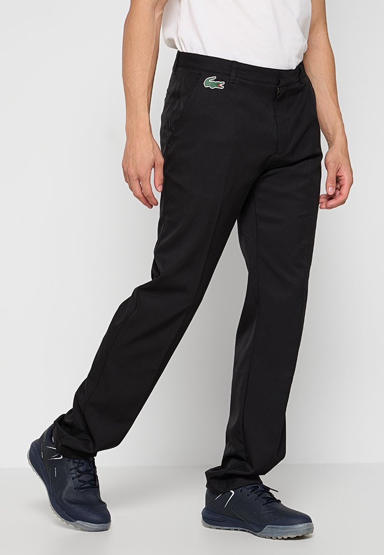 Homme portant un pantalon noir Lacoste et des baskets bleu marine, se tenant avec une jambe légèrement pliée contre un fond blanc uni.