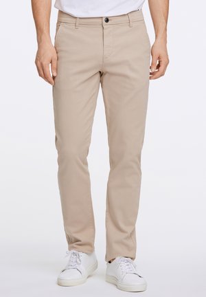 Mężczyzna w beżowych, wąskich chinosach i białych tenisówkach, stojący na tle jednolitego tła.