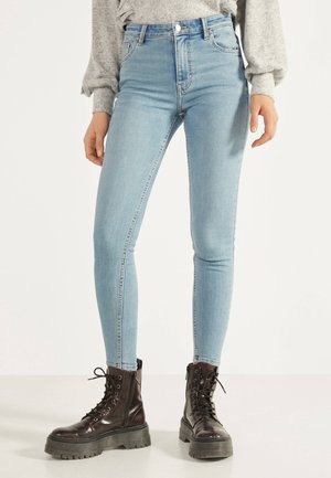 Femme portant un jean skinny bleu clair et des bottines à plateforme lacées marron foncé, debout sur un sol blanc.