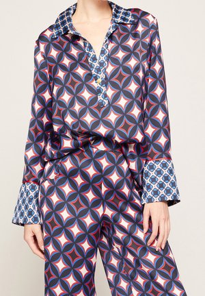 Vrouw draagt een bijpassende set van blouse en broek met een gedurfd geometrisch patroon in blauw, wit en rood, met contrasterende manchetten en kraagdetails.