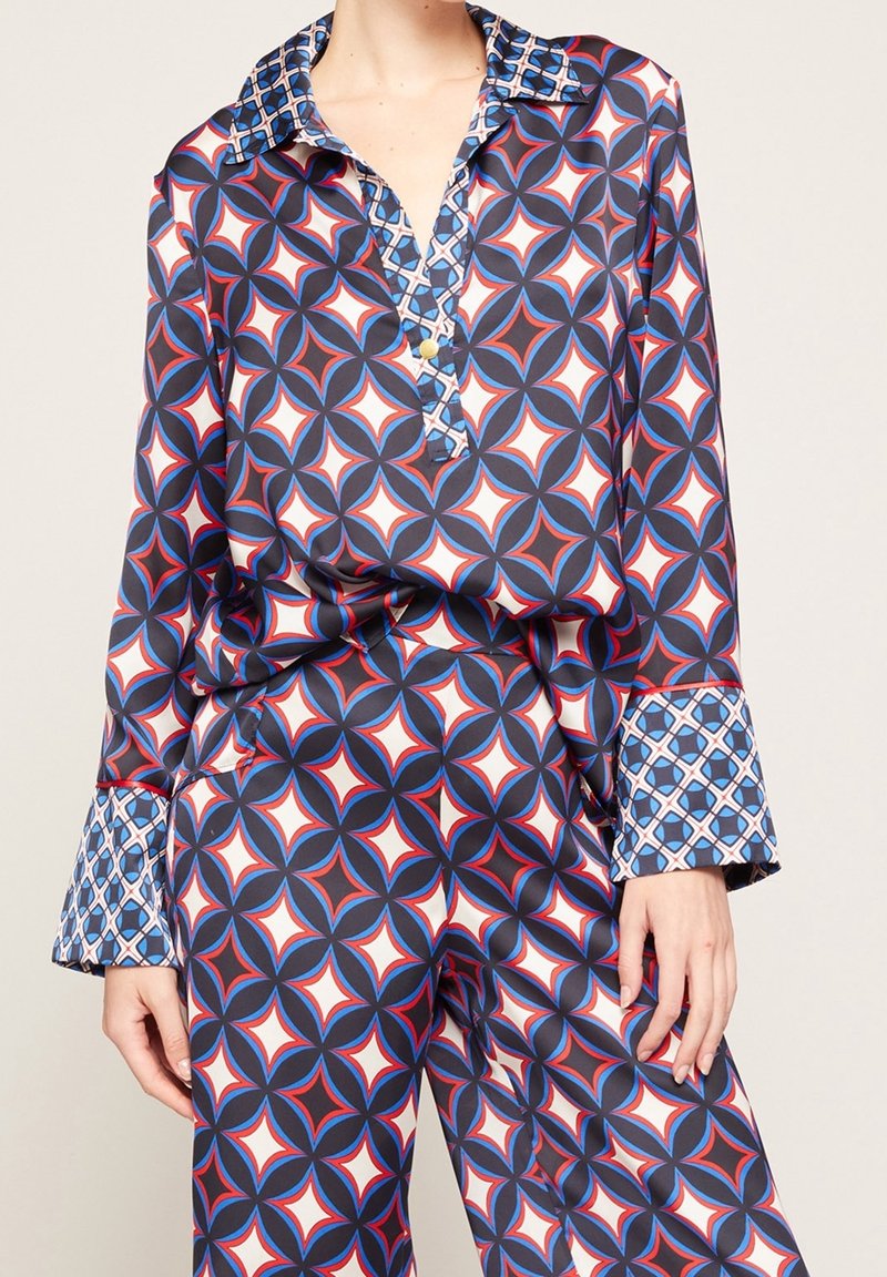 Femme portant un ensemble chemise et pantalon assorti avec un motif géométrique audacieux en bleu, blanc et rouge, avec des détails contrastants aux poignets et au col.