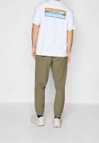 Man som bär en vit Patagonia t-shirt med berg- och våglogotyp på ryggen, olivgröna joggerbyxor och vita sneakers, står vänd bort.