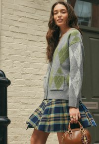 Femme portant un pull en argyle et une jupe à carreaux, tenant un sac à main marron, se tenant contre un mur en briques claires près d'une porte verte.