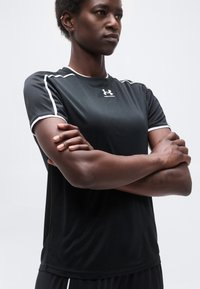 Αθλητικό άτομο με σταυρωμένα τα χέρια φορώντας μαύρο αθλητικό πουκάμισο Under Armour με λευκή περίμετρο, κοιτάζοντας εμπρός σε απλό φωτεινό φόντο.