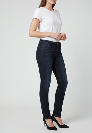 Mujer con camisa blanca de manga corta, vaqueros ajustados azul oscuro y zapatos negros de tacón alto, posando sobre un fondo liso.