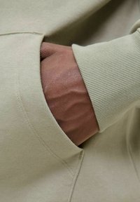 Tessuto di felpa verde chiaro con polsini a coste, presentante una mano infila in una tasca frontale. La texture è liscia, con dettagli di cucitura visibili.