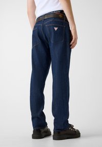 Mężczyzna ubrany w ciemnoniebieskie jeansy z tylnymi kieszeniami i naszywką z logo, brązowy pasek, czarne buty i biała koszula, stoi na jednolitym tle.