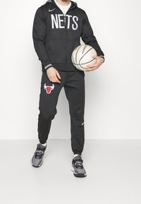Haut zippé noir Nike avec l'inscription "NETS", associé à un pantalon de survêtement noir arborant un logo rouge des Bulls. Tient un ballon de basket beige, portant des chaussures de sport grises.