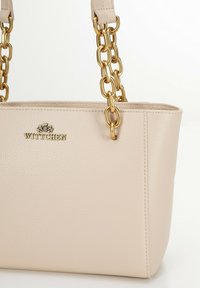 Beige leren handtas met gestructureerd oppervlak, schouderriem van goudkleurige ketting en gegraveerd logo aan de voorkant. Rechthoekige vorm.