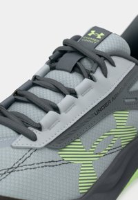 Κοντινή λήψη του γκρι παπουτσιού Under Armour Charged Commit με μαύρες κορδόνες και λογότυπο σε φανατισμένο πράσινο με λωρίδες στο πλάι.