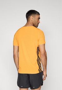 Πορτοκαλί αθλητική μπλούζα με διατρητή υφή, κοντά μανίκια και μαύρες ρίγες στα πλάγια. Φορώντας την με μαύρο σορτς, σε θέαση από πίσω.