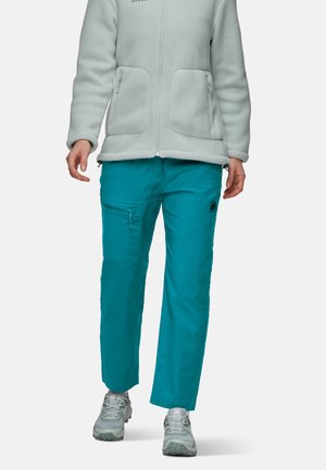 Person trägt hellgraue Fleecejacke, türkisfarbene Cargohosen mit Seitentasche und graue Wanderschuhe und geht vorwärts.